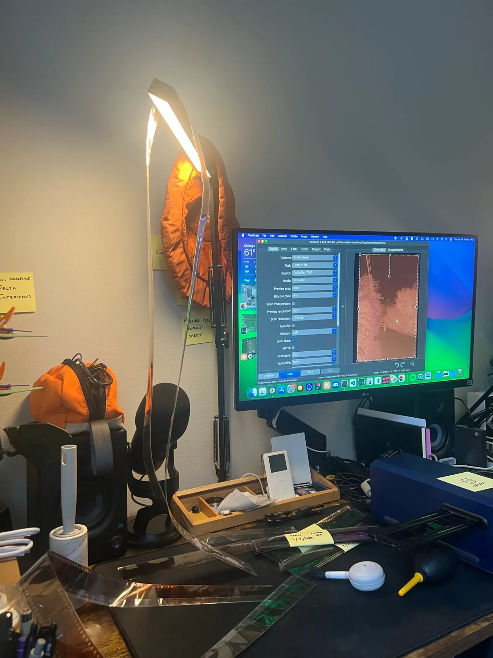 A desk lit by a lamp, with a strip of negatives hanging from it, VueScan open on a monitor showing an orange negative, and film holders, a blower and a brush next to a blue Plustek scanner.